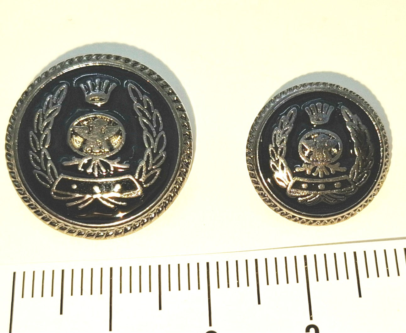 Two military-style buttons with a emblem design on a white background, accompanied by a ruler for scale.